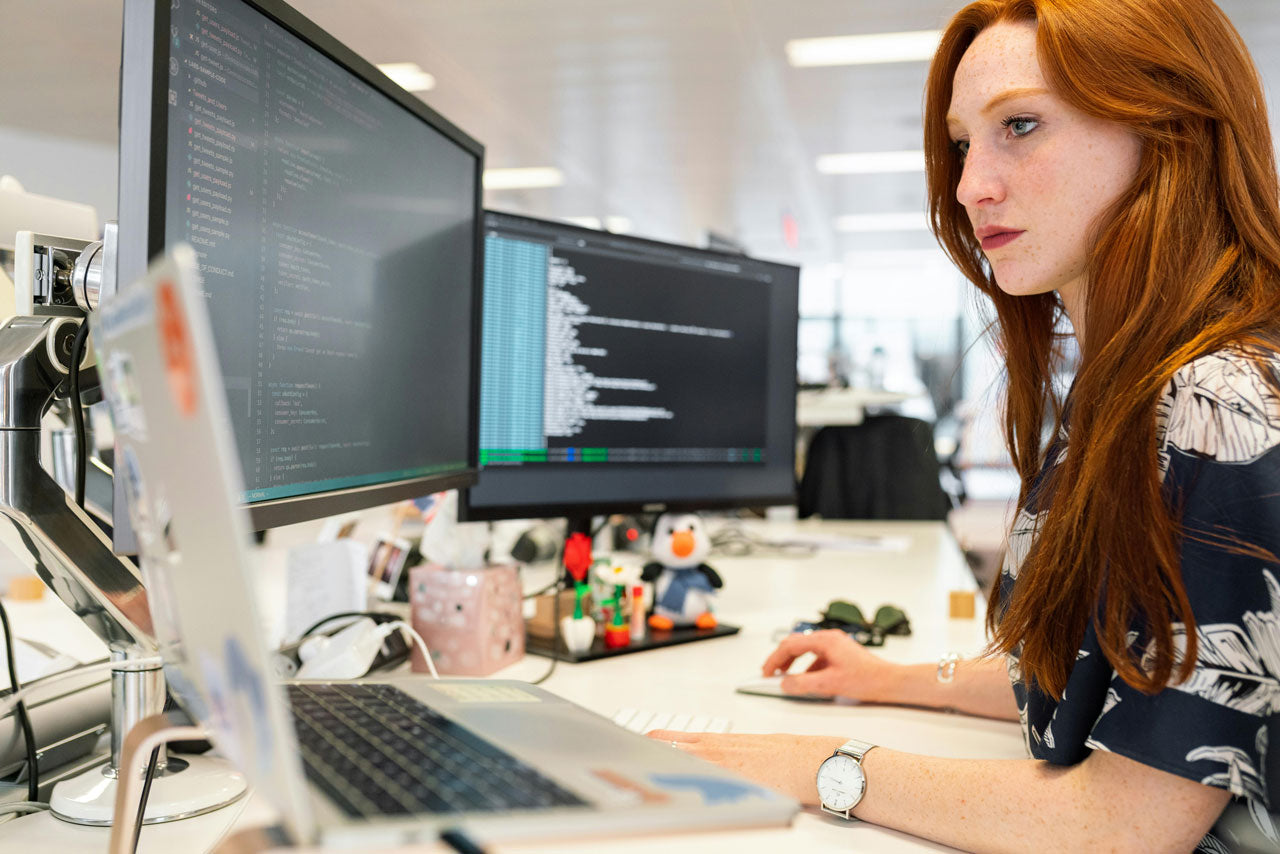 Person working at a desk with a laptop and two external monitors, focused on the screens in a modern office workspace, illustrating prolonged computer use and screen-based work.