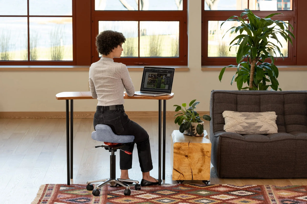 Person working at a laptop while seated on a gray QOR360 Ariel 2.0 active chair in a bright home office.